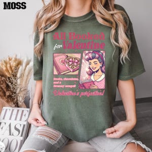 Puede incluir: Una camiseta verde con un dise&ntilde;o gr&aacute;fico rosa y blanco. El gr&aacute;fico presenta a una mujer leyendo un libro y una caja de chocolates. El texto en la camiseta dice "All Booked for Valentine. Books, chocolates, and a dreamy escape! Valentine's perfection!"
