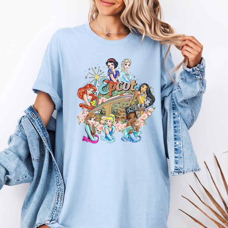 May include: Light blue t-shirt featuring a graphic of Disney princesses in front of the Epcot logo. The princesses include Ariel, Snow White, Cinderella, Belle, and Elsa.