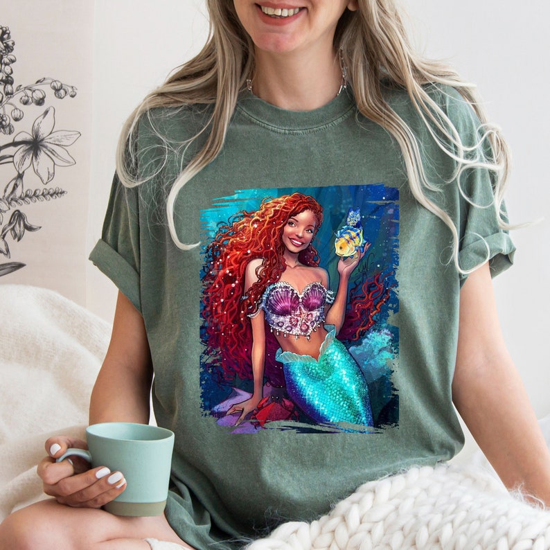 May include: A green t-shirt featuring a graphic print of a red-haired mermaid holding a yellow and blue fish. The mermaid is wearing a pink and purple seashell bra and a turquoise tail.