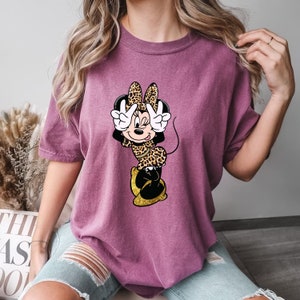 K&ouml;nnte beinhalten: Mauvefarbenes T-Shirt mit einer Cartoon-Figur von Minnie Mouse in einem Leopardenmuster-Outfit. Minnie macht mit beiden H&auml;nden ein Friedenszeichen. Ihre Schuhe sind goldfarben glitzernd. Das Shirt hat kurze &Auml;rmel und eine lockere Passform.