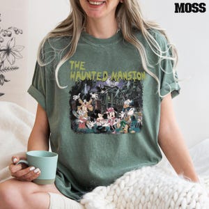 May include: A green t-shirt with a vintage-style graphic of Mickey Mouse and friends in front of a haunted mansion. The text "The Haunted Mansion" is printed in yellow above the image.