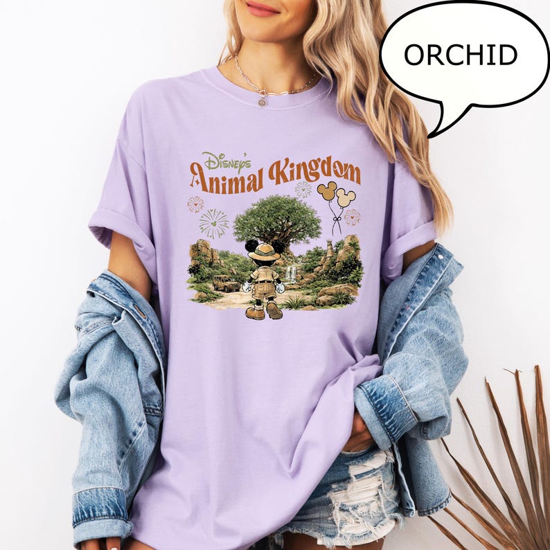 May include: Orchid-colored t-shirt featuring the text "Disney's Animal Kingdom" with a graphic of Mickey Mouse in safari attire in front of a tree. The shirt has rolled-up sleeves and is paired with a denim jacket.