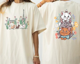 May include: Two white t-shirts with different designs. The front of the shirt features a cartoon illustration of three cats in front of the Eiffel Tower. The back of the shirt features a cartoon illustration of three cats with flowers.