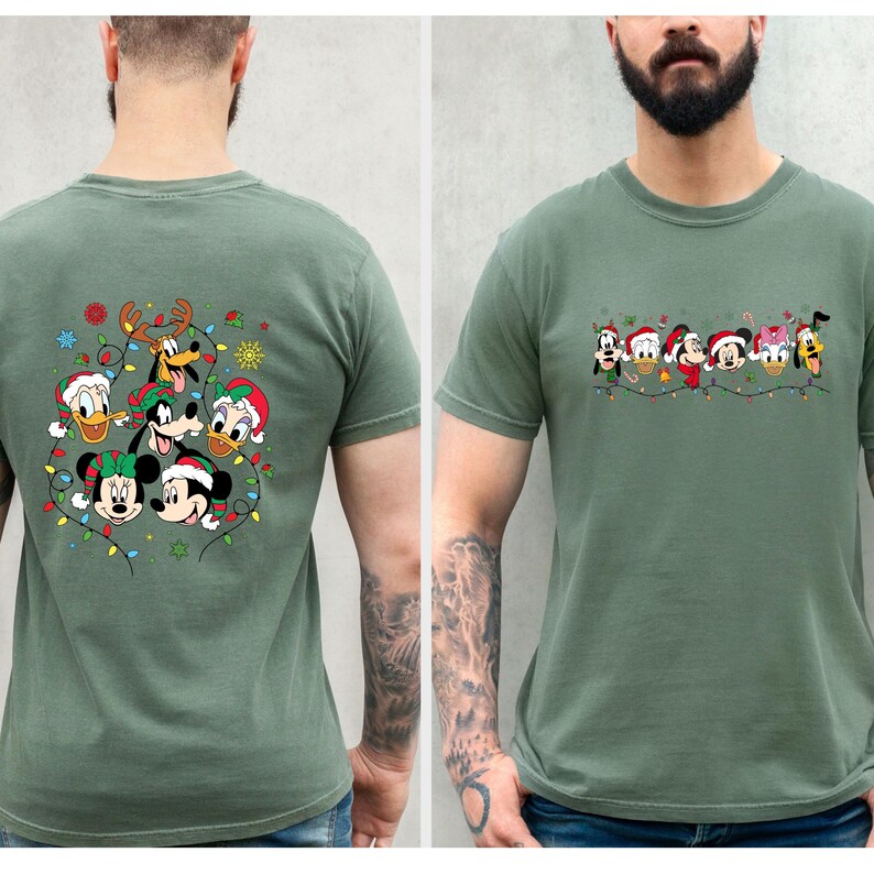 May include: Sage green t-shirt featuring a festive design with Disney characters wearing Santa hats and Christmas lights. The back of the shirt showcases Mickey, Minnie, Donald, and Goofy. The front displays a row of Disney characters.