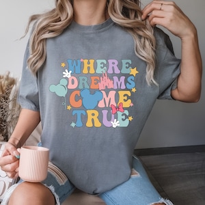 May include: A gray t-shirt with the text "Where Dreams Come True" in colorful letters. The text is surrounded by cartoon images of Mickey Mouse ears, a castle, stars, and a bow.