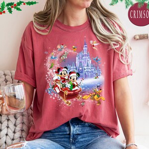 Puede incluir: Una camiseta roja con un dise&ntilde;o navide&ntilde;o que presenta a Mickey Mouse y Minnie Mouse montando en un trineo con un castillo en el fondo. El dise&ntilde;o tambi&eacute;n incluye otros personajes de Disney como Pluto y Campanilla.