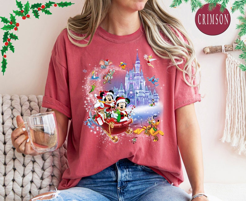 Puede incluir: Una camiseta roja con un dise&ntilde;o navide&ntilde;o que presenta a Mickey Mouse y Minnie Mouse montando en un trineo con un castillo en el fondo. El dise&ntilde;o tambi&eacute;n incluye otros personajes de Disney como Pluto y Campanilla.