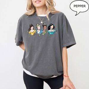 May include: Dark gray t-shirt featuring a watercolor illustration of four female figures with various hairstyles and dresses. The word "PEPPER" is in a speech bubble. The shirt has short sleeves and a white undershirt.