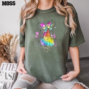 Puede incluir: Una camiseta verde con una ilustraci&oacute;n de acuarela arco&iacute;ris de un castillo con orejas de Mickey Mouse y globos.