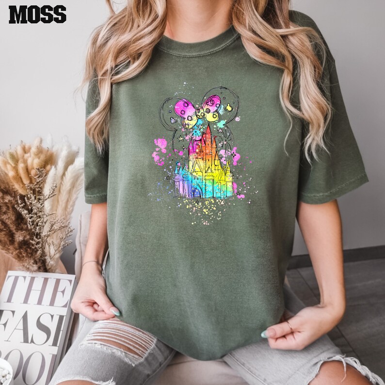 Puede incluir: Una camiseta verde con una ilustraci&oacute;n de acuarela arco&iacute;ris de un castillo con orejas de Mickey Mouse y globos.