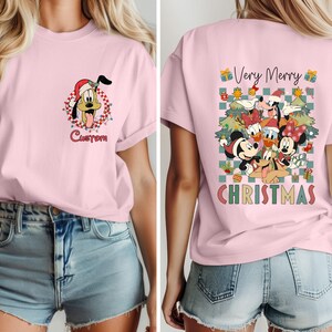 Pode incluir: Camiseta rosa claro com um desenho animado de Pluto usando um gorro de Papai Noel e luzes de Natal. A palavra "Custom" est&aacute; abaixo da imagem. A parte de tr&aacute;s da camisa tem um design "Very Merry Christmas" com Mickey, Minnie, Donald e Pateta.