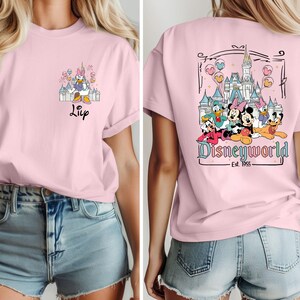 May include: Pink t-shirt with a graphic of Disney characters in front of the Cinderella Castle. The back of the shirt has a graphic of Mickey Mouse, Minnie Mouse, Donald Duck, Goofy, Pluto, and Daisy Duck in front of the Cinderella Castle with the text "Disneyworld - Est. 1955".