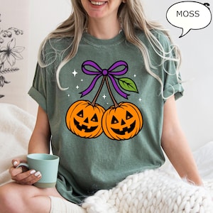 May include: A moss green t-shirt featuring a Halloween-themed graphic. The graphic depicts two orange jack-o'-lanterns with smiling faces, connected by a purple bow and green leaf. The word "MOSS" is in a speech bubble.