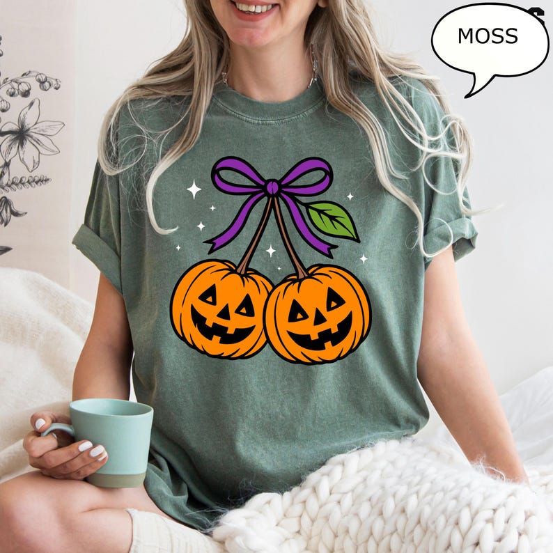 May include: A moss green t-shirt featuring a Halloween-themed graphic. The graphic depicts two orange jack-o'-lanterns with smiling faces, connected by a purple bow and green leaf. The word "MOSS" is in a speech bubble.