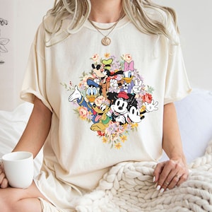 May include: A cream coloured t-shirt with a graphic print of Mickey Mouse, Minnie Mouse, Donald Duck, Goofy, and Pluto surrounded by colourful flowers.