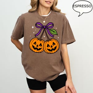 May include: Brown t-shirt featuring a Halloween-themed graphic. The design includes two orange jack-o'-lanterns connected by a purple bow and green leaf. The word "ESPRESSO" is in a speech bubble. The shirt is a casual, oversized fit.