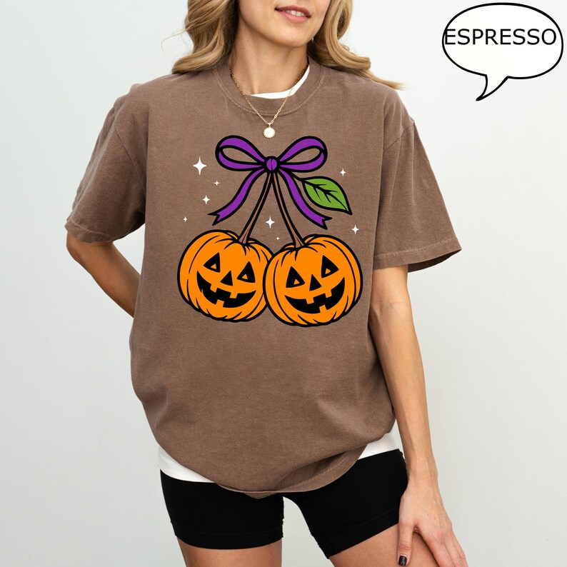 May include: Brown t-shirt featuring a Halloween-themed graphic. The design includes two orange jack-o'-lanterns connected by a purple bow and green leaf. The word "ESPRESSO" is in a speech bubble. The shirt is a casual, oversized fit.