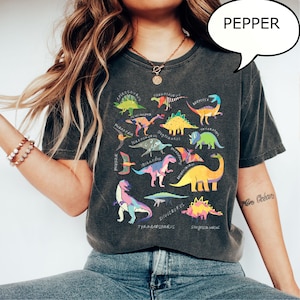 May include: Dark gray t-shirt featuring a colorful dinosaur graphic with various species names. The design includes a variety of dinosaurs in shades of green, yellow, blue, and pink. The word "PEPPER" is in a speech bubble.