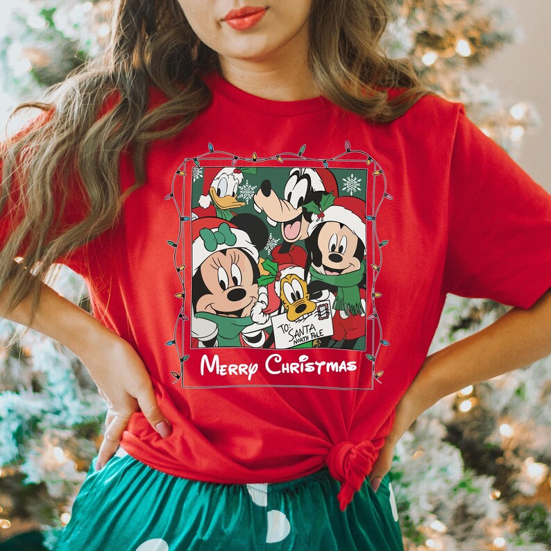May include: Red t-shirt with a Christmas-themed graphic featuring Mickey Mouse, Minnie Mouse, Donald Duck, Goofy, and Pluto. The graphic is surrounded by a string of Christmas lights and the text "Merry Christmas".