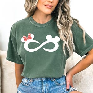 K&ouml;nnte beinhalten: Ein gr&uuml;nes T-Shirt mit einem wei&szlig;en Unendlichkeitszeichen und einer roten Schleife mit wei&szlig;en Punkten auf einer Seite. Das Unendlichkeitszeichen hat auf jeder Seite ein Mickey-Maus-Ohr.