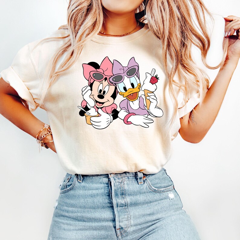 Puede incluir: Una camiseta blanca con una imagen de dibujos animados de Minnie Mouse y Daisy Duck con gafas de sol y sosteniendo conos de helado. La imagen es en rosa, morado y amarillo.