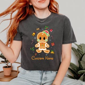 May include: A dark gray cropped t-shirt with a gingerbread man wearing a Santa hat and surrounded by Christmas decorations. The text "Custom Name" is printed below the gingerbread man.