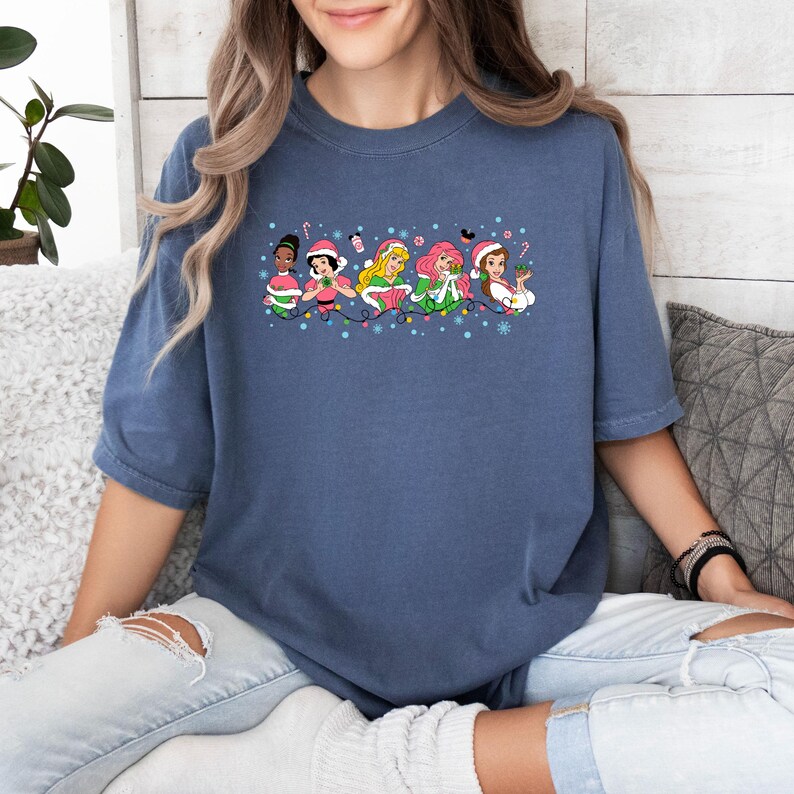 Puede incluir: Una camiseta azul con un gr&aacute;fico de seis princesas Disney con gorros de Pap&aacute; Noel y sosteniendo art&iacute;culos navide&ntilde;os. Las princesas est&aacute;n alineadas en una fila con luces navide&ntilde;as y copos de nieve a su alrededor.
