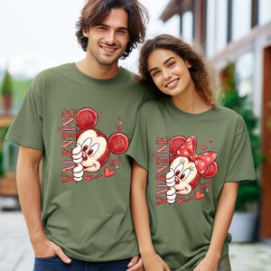 Puede incluir: Dos camisetas verde oliva con personajes de dibujos animados. Una presenta a Mickey Mouse, la otra a Minnie Mouse, ambas con la palabra "VALENTINE" en rojo. Corazones y flores complementan el dise&ntilde;o, perfecto para San Valent&iacute;n.