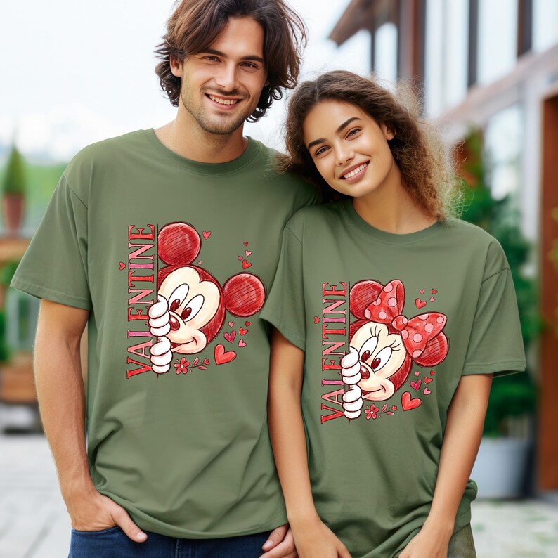 Puede incluir: Dos camisetas verde oliva con personajes de dibujos animados. Una presenta a Mickey Mouse, la otra a Minnie Mouse, ambas con la palabra "VALENTINE" en rojo. Corazones y flores complementan el dise&ntilde;o, perfecto para San Valent&iacute;n.