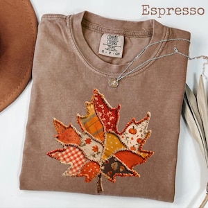 May include: An espresso-colored t-shirt featuring a large, central maple leaf graphic composed of various fabric patches in orange, red, and yellow tones with plaid, floral, and pumpkin patterns. The shirt has a crew neckline and a Comfort Colors label.
