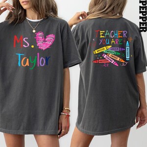 May include: Dark gray t-shirt with the text "Ms. Taylor" in colorful letters and a pink heart on the front. The back of the shirt says "Teacher You Are" with crayon graphics and positive words.