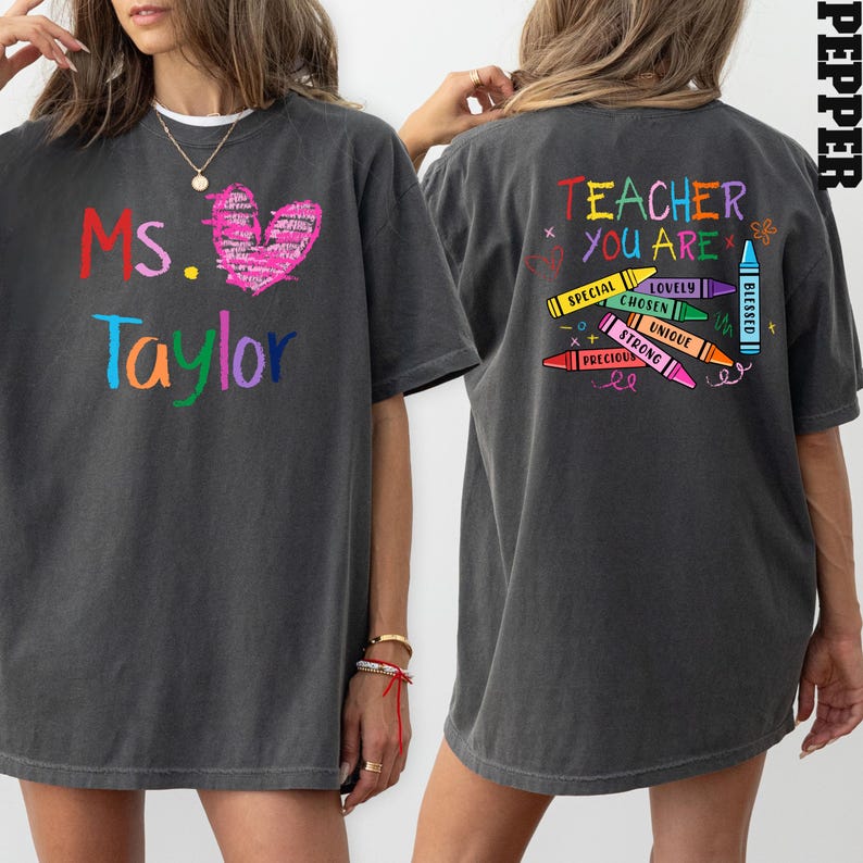 May include: Dark gray t-shirt with the text "Ms. Taylor" in colorful letters and a pink heart on the front. The back of the shirt says "Teacher You Are" with crayon graphics and positive words.