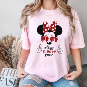 May include: Pink t-shirt with a graphic of Mickey Mouse wearing sunglasses with the text "First Disney Trip" on the front.