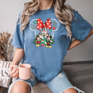 Puede incluir: Una camiseta azul con un lazo rojo y blanco, un &aacute;rbol de Navidad, Mickey Mouse y Minnie Mouse, y un castillo en el fondo.