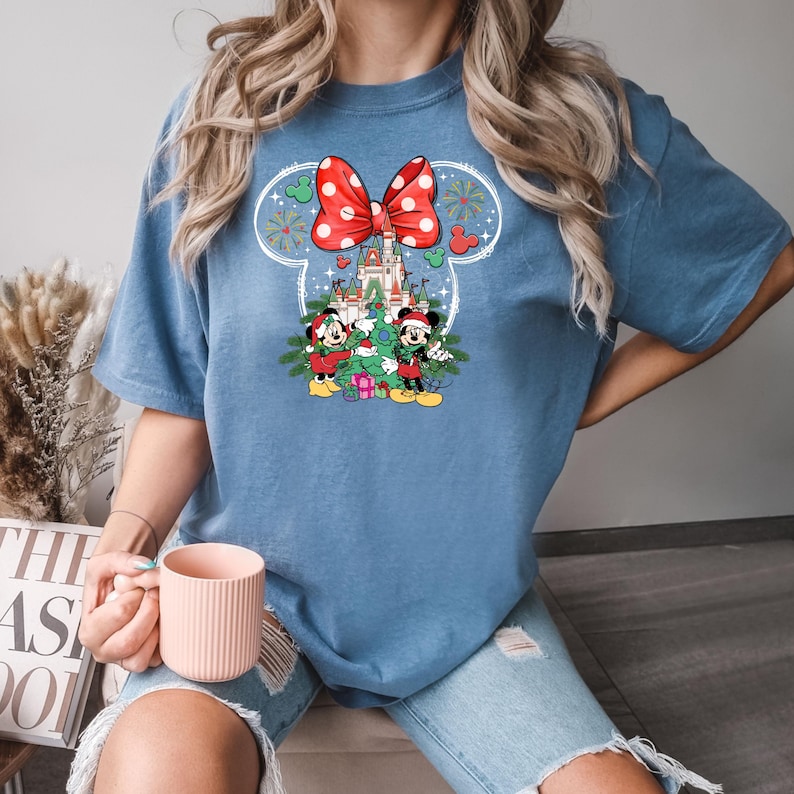 Puede incluir: Una camiseta azul con un lazo rojo y blanco, un &aacute;rbol de Navidad, Mickey Mouse y Minnie Mouse, y un castillo en el fondo.