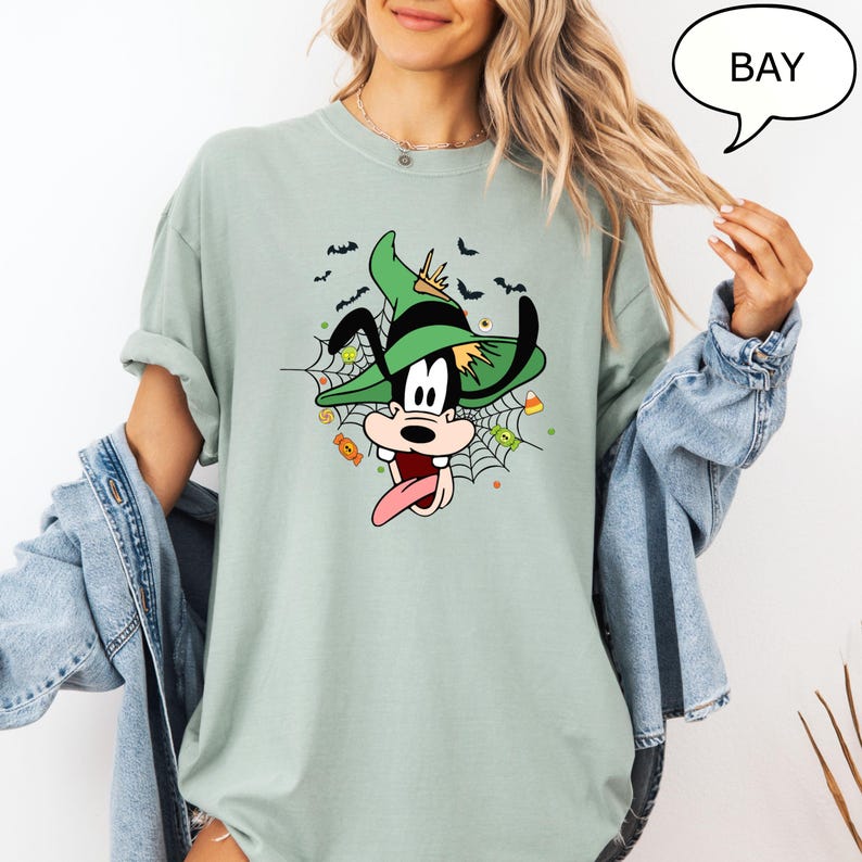 Może przedstawiać: Jasnozielona koszulka z postacią z kresk&oacute;wki w zielonym kapeluszu czarownicy, otoczona pajęczyną i słodyczami na Halloween. Postać wystawia język. Słowo "BAY" znajduje się w dymku.