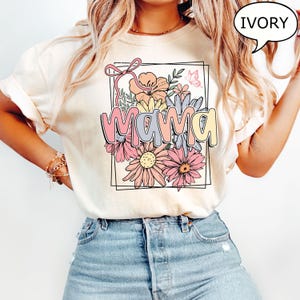 May include: A white t-shirt with a floral design and the word "mama" in a colorful font. The shirt is ivory in color.