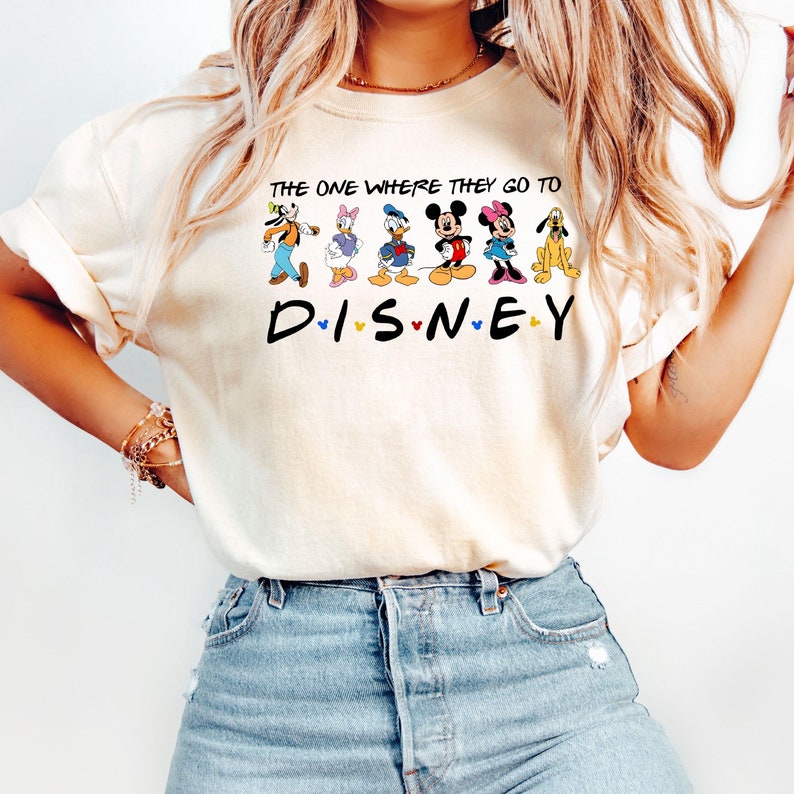 May include: Cream-colored t-shirt featuring the text "THE ONE WHERE THEY GO TO DISNEY" with Disney characters. The design includes Goofy, Daisy Duck, Donald Duck, Mickey Mouse, Minnie Mouse, and Pluto.