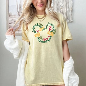 K&ouml;nnte beinhalten: Ein gelbes T-Shirt mit einem gr&uuml;nen floralen Mickey Mouse-Design. Das Design enth&auml;lt rote Herzen und gelbe Blumen.