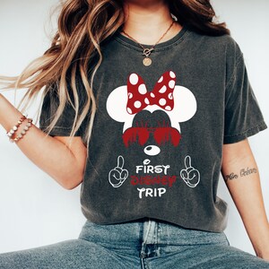 May include: A dark gray t-shirt with a white silhouette of Minnie Mouse wearing sunglasses. The bow is red with white polka dots. The text "First Disney Trip" is in red with white outlines. The text is surrounded by two white hands with index fingers pointing up.