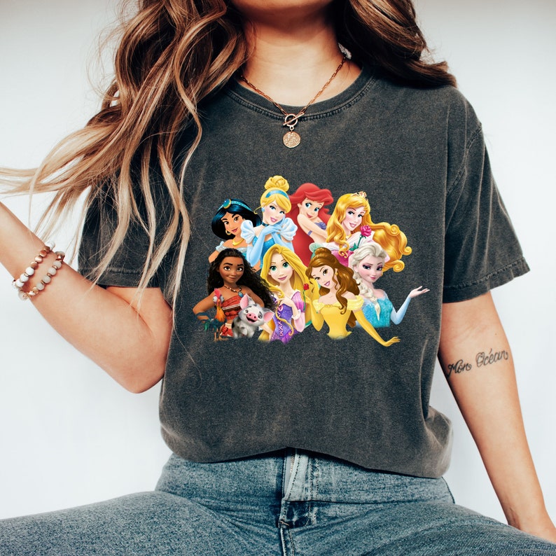 Puede incluir: Una camiseta gris oscuro con un estampado gr&aacute;fico de princesas Disney, incluyendo Cenicienta, Ariel, Bella, Jasmine y Moana. Las princesas est&aacute;n todas sonriendo y mirando al espectador.