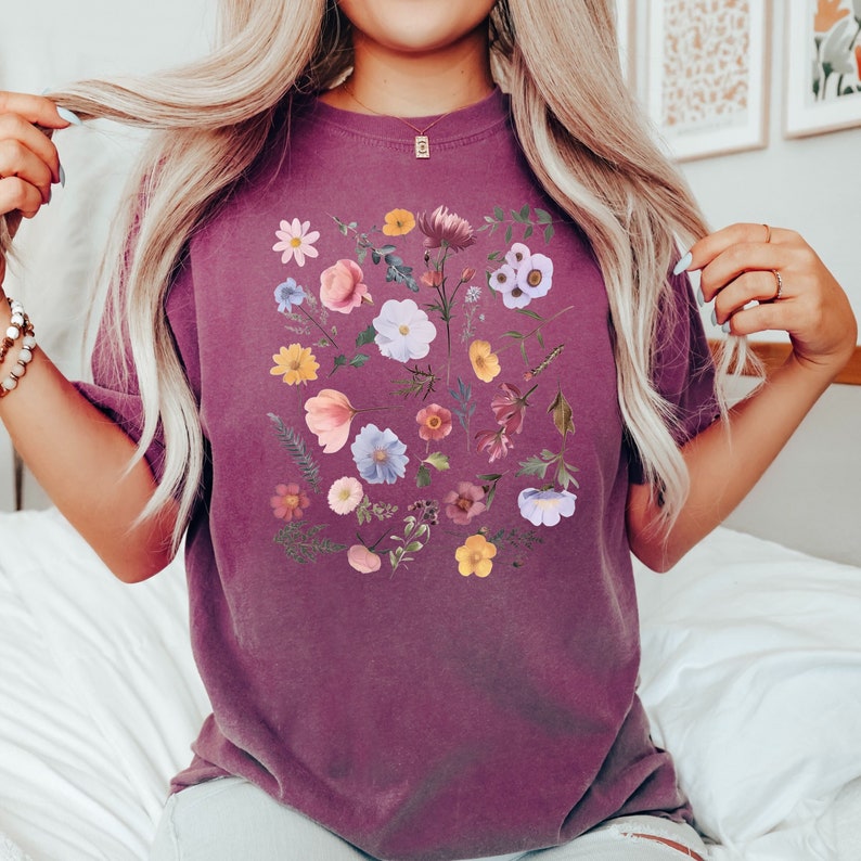 May include: A purple t-shirt with a floral print featuring a variety of flowers in shades of pink, purple, blue, and yellow.