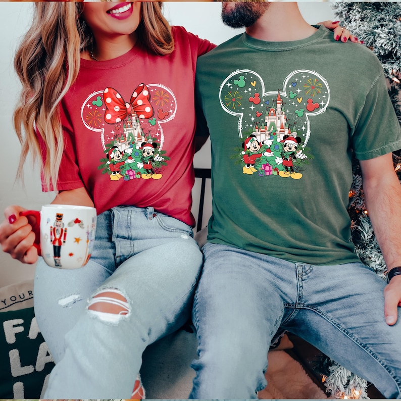 Puede incluir: Una pareja lleva camisetas a juego con tem&aacute;tica navide&ntilde;a. La camiseta de la mujer es roja con una cabeza de Mickey Mouse y un lazo. La camiseta del hombre es verde con una cabeza de Mickey Mouse y un &aacute;rbol de Navidad. Ambas camisetas tienen un castillo y fuegos artificiales.