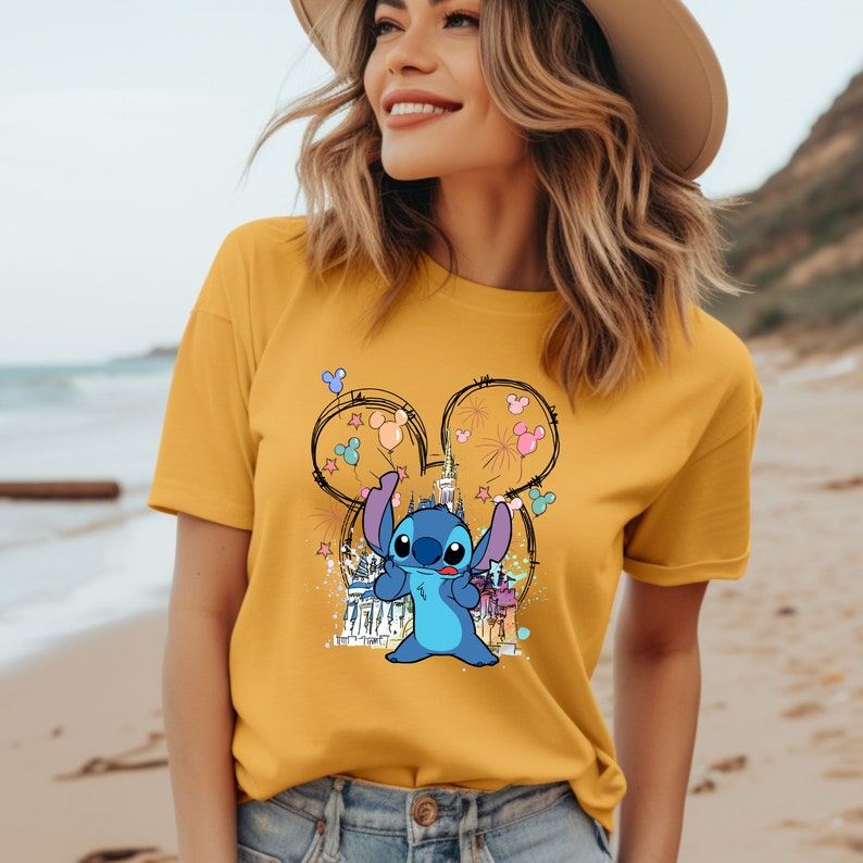 May include: A yellow t-shirt with a blue Stitch character holding balloons in front of a cartoon drawing of a castle. The balloons are pink, purple, blue, green, and yellow. The castle is outlined in black and has a Mickey Mouse ear shape.