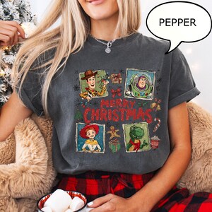 May include: Dark gray t-shirt featuring a "Merry Christmas" design with cartoon characters. The design includes Woody, Buzz Lightyear, Jessie, and Rex, with Christmas-themed elements. The word "Pepper" is in a speech bubble.