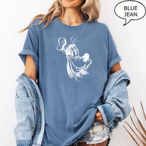 Puede incluir: Una camiseta azul vaquero con un dibujo de contorno blanco de un perro de dibujos animados con una gran sonrisa. El perro lleva un sombrero.