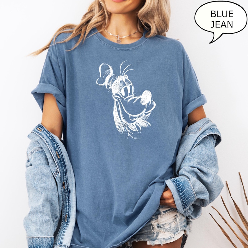 Puede incluir: Una camiseta azul vaquero con un dibujo de contorno blanco de un perro de dibujos animados con una gran sonrisa. El perro lleva un sombrero.