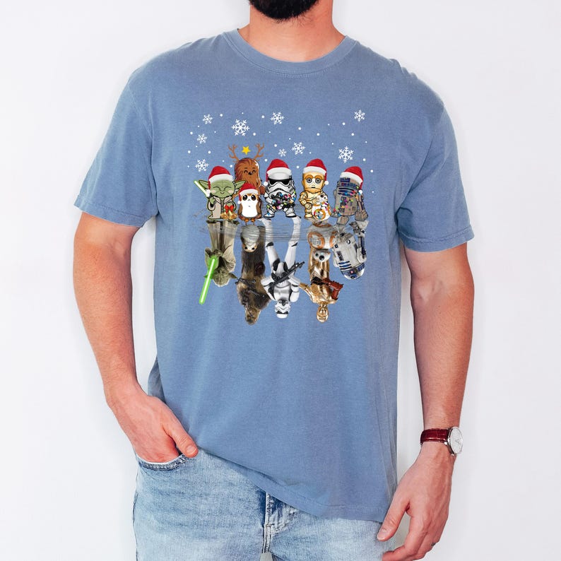 Puede incluir: Una camiseta azul con un dise&ntilde;o de Star Wars con tem&aacute;tica navide&ntilde;a. El dise&ntilde;o presenta varios personajes de Star Wars con gorros de Pap&aacute; Noel y reflejados en un charco de agua. Los personajes incluyen a Yoda, Chewbacca, Darth Vader, un Stormtrooper, R2-D2 y C-3PO.