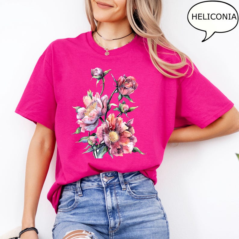 Pu&ograve; includere: Una maglietta magenta con un design floreale ad acquerello. Il design presenta fiori rosa, pesca e gialli. La maglietta ha maniche corte e girocollo. La parola "HELICONIA" &egrave; in un fumetto.