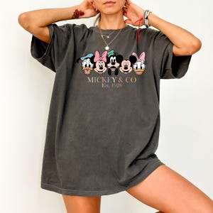 May include: A dark gray oversized t-shirt with a graphic print of Mickey Mouse and his friends. The print features Mickey Mouse, Minnie Mouse, Donald Duck, Goofy, and Pluto. The text below the characters reads "MICKEY & CO Est. 1928".